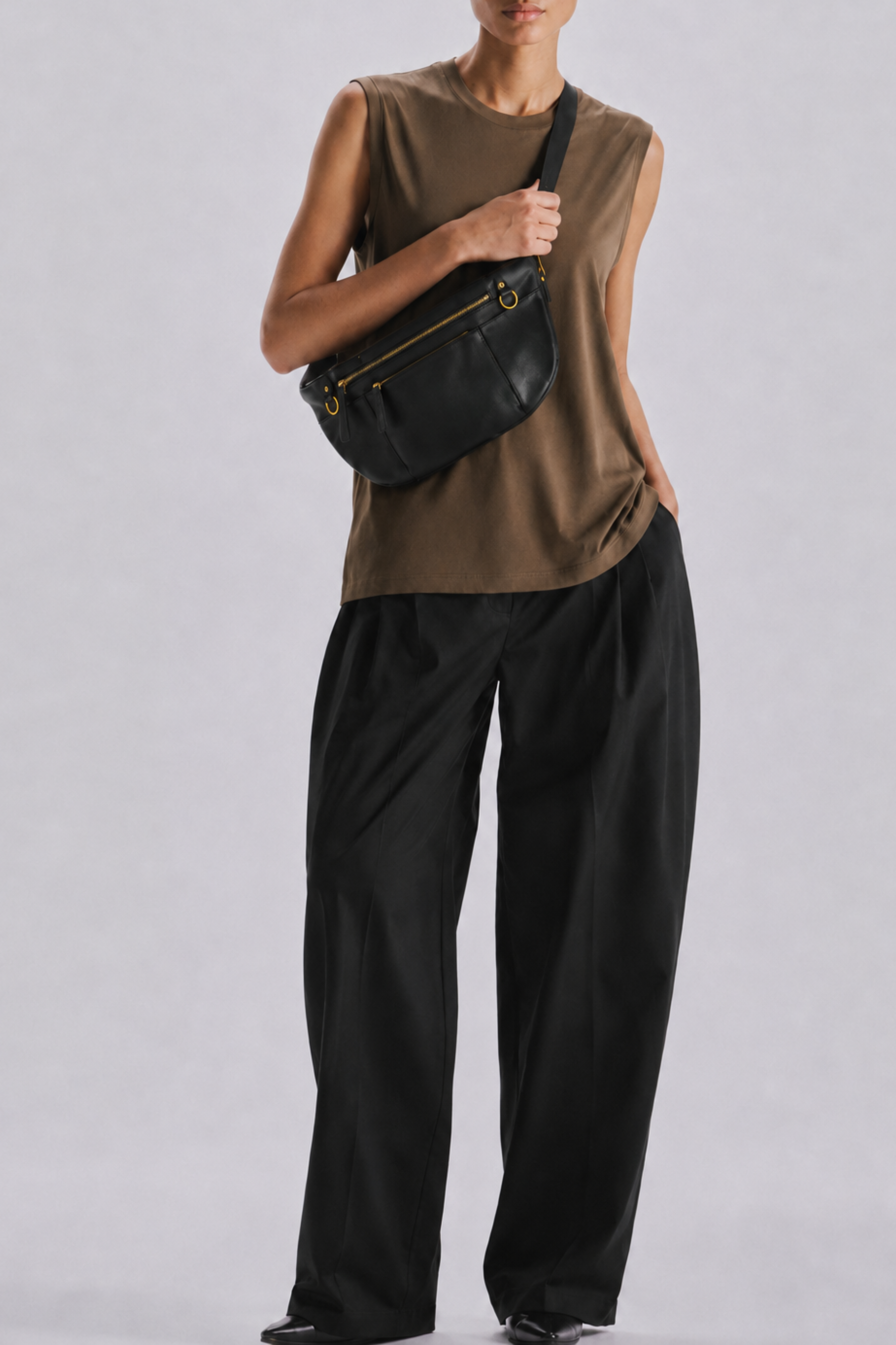 A model wearing a sleeveless brown top, black wide-leg pants, and a black leather crossbody bag with gold hardware, standing against a light gray studio background.