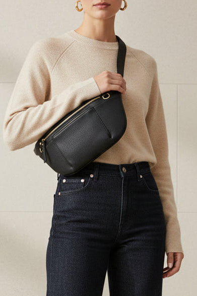 Black leather commuter sling bag worn crossbody, featuring an adjustable strap, zip compartments, and gold hardware, styled as a hands-free everyday bag for work, travel, and daily use.