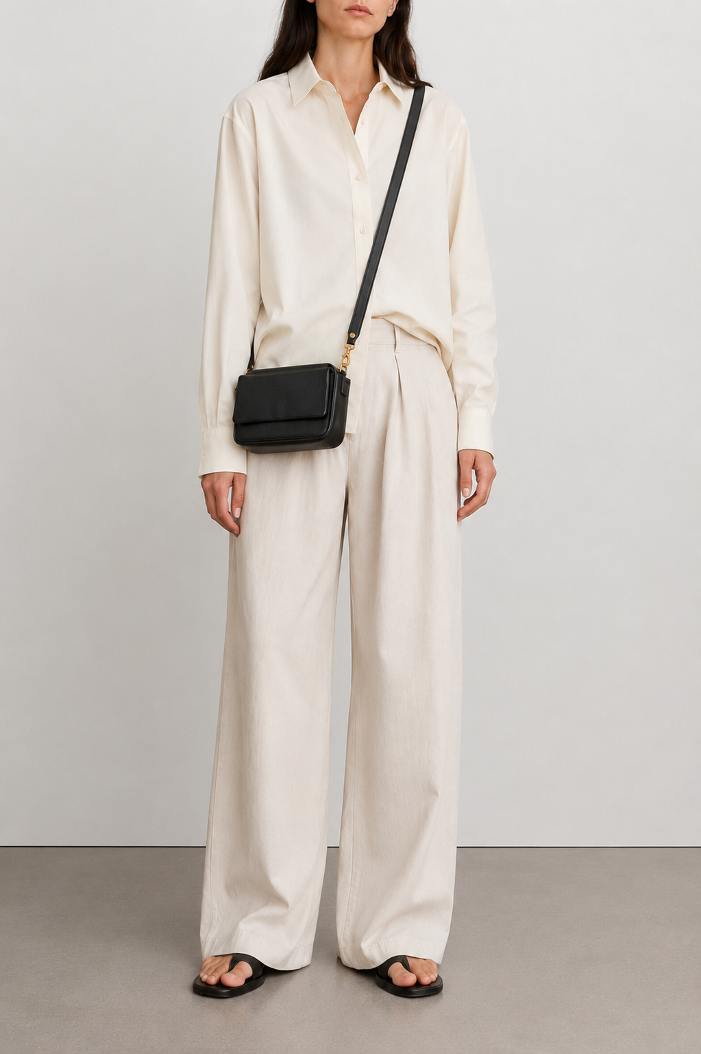 A model wearing a cream-colored long-sleeve button-up shirt and matching wide-leg trousers, styled with a small black crossbody bag and black sandals, set against a neutral studio background.