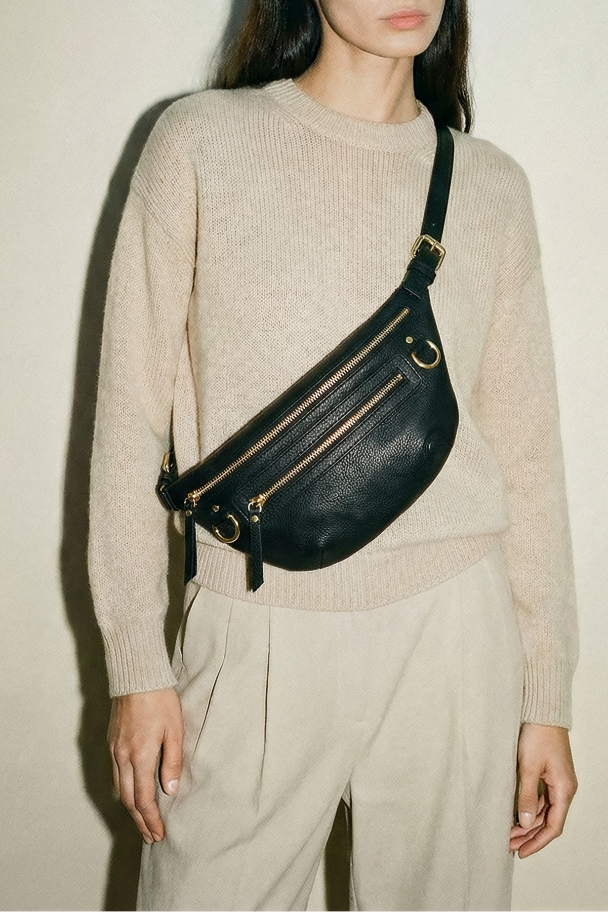 Woman wearing a beige sweater and light pants with a black leather fanny pack against a plain background
